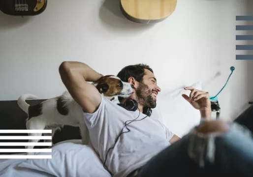 Man lying on a bed with headphones, laughing as a small dog licks his face.