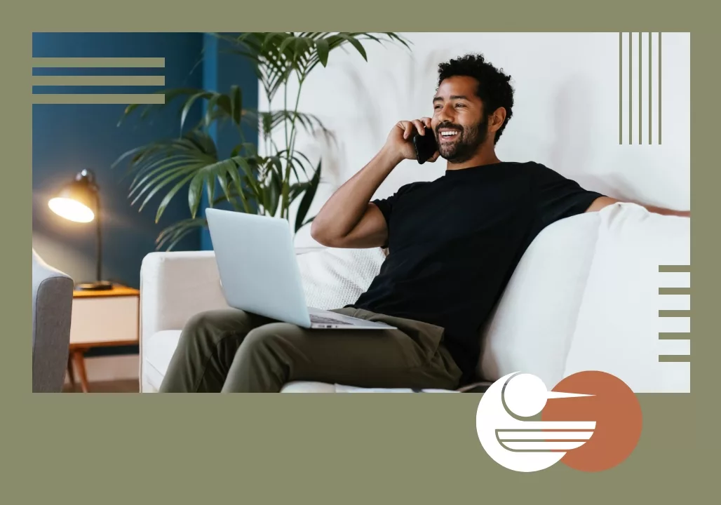 Man smiling on a white sofa with a laptop, talking on the phone, surrounded by plants and a lamp in a cozy setting.