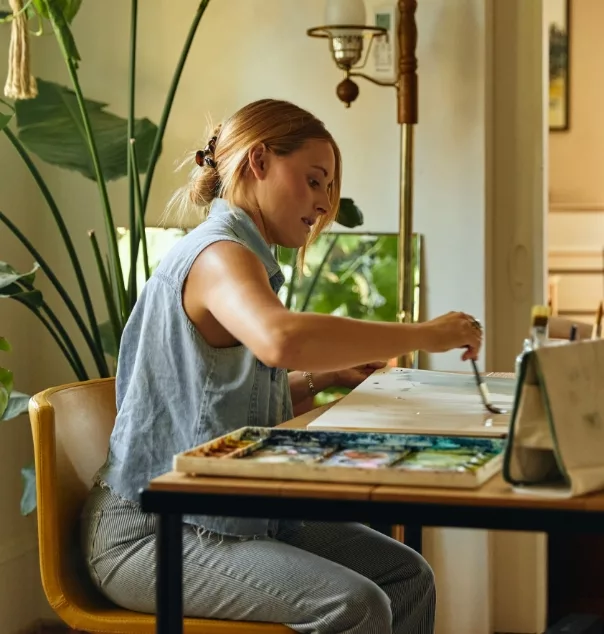 Woman painting at a desk in a bright, plant-filled room with natural light.