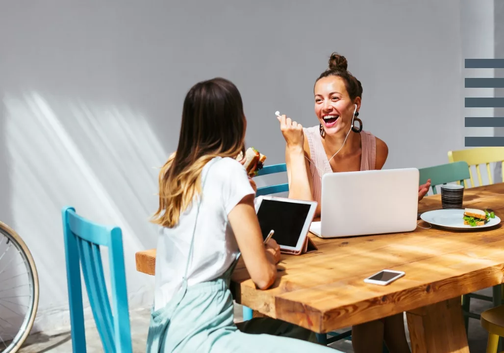 Two women laughing and working on a wooden table with a laptop and tablet, surrounded by colorful chairs.