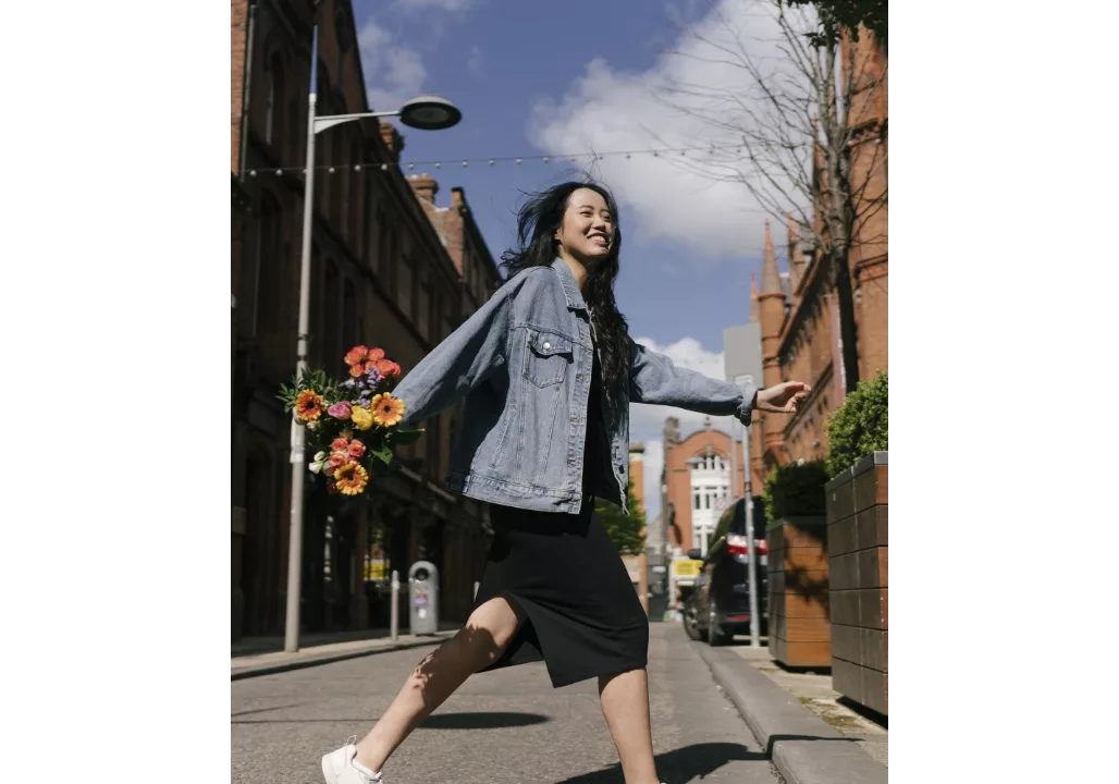 Woman walking joyfully on a street holding a bouquet of flowers, wearing a denim jacket and black dress.