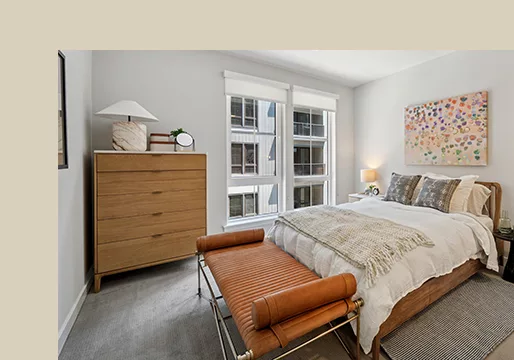 A photo of a bedroom with white walls and a gray carpet with a bed, orange settee, dresser, and large windows.
