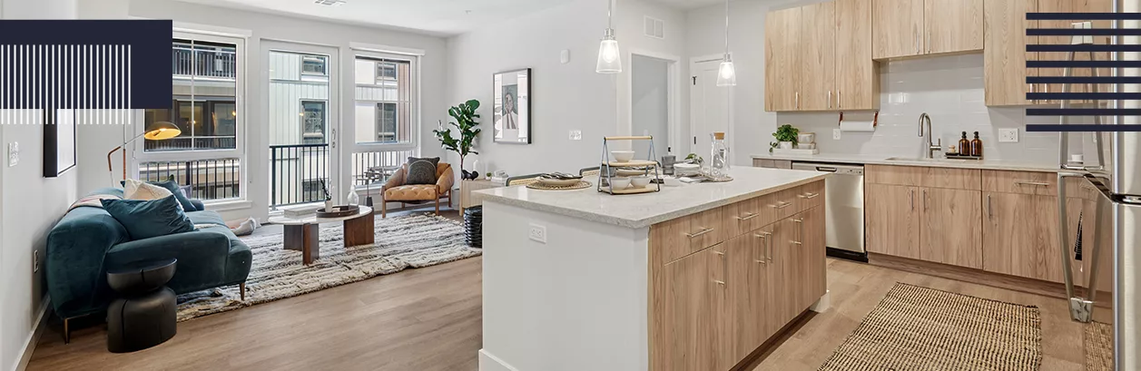 An open-concept living apartment with white walls, light-color wood cabinets and flooring, and large windows.